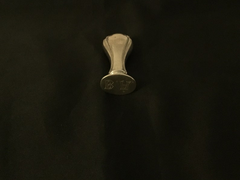 Op de afbeelding: Een zilveren zegel met een afgeronde bovenkant en een handvat. De zegel heeft de initialen "B.H." gegraveerd op de onderkant.