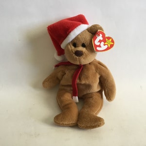 May include: A brown teddy bear wearing a red Santa hat with a white trim and a red scarf. The bear has a red heart-shaped tag with the words "Ty" and "Beanie Babies" on it.