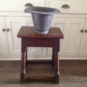 Puede incluir: Un macetero de metal plateado con forma de sombrero se encuentra sobre un taburete de madera marrón oscuro con patas torneadas. El taburete tiene una tapa cuadrada y está delante de un armario blanco con dos puertas.