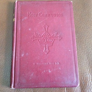 Puede incluir: Un libro de color granate titulado "Holy Communion" con un diseño de cruz en relieve en la portada. El nombre del autor, W. Walsham How D.D., está impreso en la parte inferior. La cubierta del libro muestra signos de desgaste.