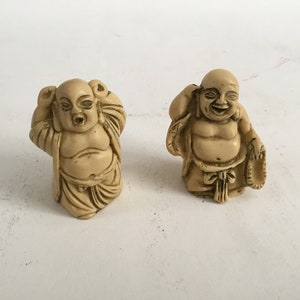Puede incluir: Dos pequeñas figuritas de color crema del Buda sonriente. Un Buda sostiene un pequeño tambor, mientras que el otro se lleva la mano a la oreja.