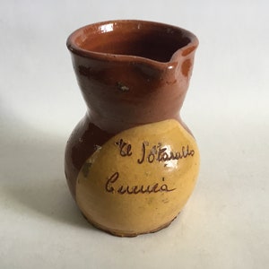 Op de afbeelding: Een bruine en gele keramische kan met een rond lichaam en een kleine tuit. De kan heeft een gele cirkel aan de zijkant met de tekst "El Jotasullo Cuenca" in bruine letters.