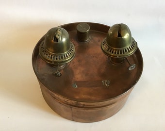 May include: A copper oil lamp with two brass burners. The lamp has a round base and a cylindrical body. The burners are attached to the top of the lamp and have a decorative design.
