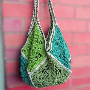 Może przedstawiać: Ręcznie robiona torba typu tote z szydełka w odcieniach zieleni i turkusu, z geometrycznym ażurowym wzorem. Torba ma solidny pasek i wisi na czerwonej ceglanej ścianie. Stylowy i ekologiczny dodatek.