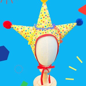May include: A yellow jester hat featuring a colorful polka dot pattern, three pointed cones topped with red, yellow, and blue pom-poms, a red fabric trim around the face opening, and a red ribbon tie closure under the chin area.