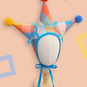 Può includere: Un cappello da giullare a più punte caratterizzato da un colorato motivo a quadri nei toni dell'arancione, del blu e del giallo. Il cappello è decorato con un pon-pon arancione brillante sulla punta, uno giallo su un lato e uno blu, con bordo in pizzo blu.