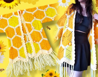 May include: A crocheted cardigan with a honeycomb pattern in yellow and white. The cardigan has a fringe trim and is worn by a person with long dark hair.