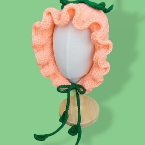 May include: A peach-coloured crocheted hat with green crocheted leaves and a bow. The hat is on a white mannequin head.