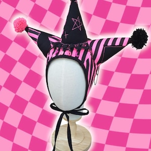 May include: A black and pink striped jester hat with a star and cross design. The hat has three pom-poms, a black ribbon tie, and a decorative trim. The hat is displayed on a mannequin head against a pink checkered background.