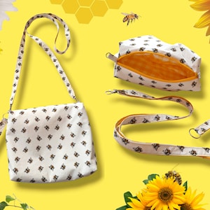 May include: A white crossbody bag, cosmetic bag, lanyard, and wristlet with a repeating pattern of bees. The bags have a yellow interior. The background is yellow with sunflowers and bees.