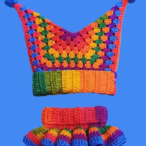 Könnte beinhalten: Ein farbenfrohes, handgefertigtes Häkelhut- und passendes Rüschenkragen-Set. Der Hut ist eine Mischung aus Regenbogenfarben mit einer spitzen Spitze und blauen Pompons. Der Kragen hat ein ähnliches Regenbogenmuster mit einem Rüschenrand.