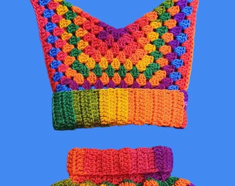 May include: A colorful, handmade crochet hat and matching ruffled collar set. The hat is a vibrant mix of rainbow colors with a pointed top and blue pom-poms. The collar features a similar rainbow pattern with a ruffled edge, perfect for a costume or playful outfit.