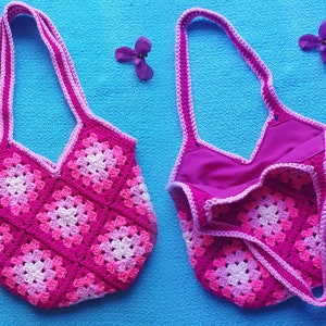 May include: A pink and white crocheted tote bag with a pink lining. The bag is made of granny squares and has a wide, flat bottom.