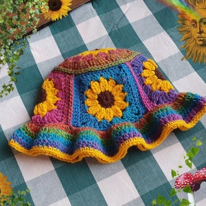 May include: A colorful crocheted sun hat with a rainbow trim and a flower design. The hat is made of yarn in shades of pink, blue, green, and yellow.