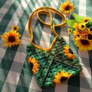 May include: A green and yellow crocheted bag with sunflower designs. The bag has a long strap and is made with a granny square stitch.
