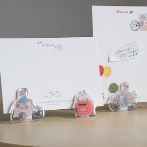 May include: Three acrylic postcard holders featuring illustrations of a cat riding a red mailbox, a person riding a bicycle, and a person driving a car. The holders are on a wooden surface. The text "Postcard" is visible on the white postcards.