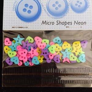 Puede incluir: Una bolsa de botones de plástico de colores neón en forma de corazones y estrellas. La bolsa está etiquetada con "Dress It Up" y "Micro Shapes Neon".