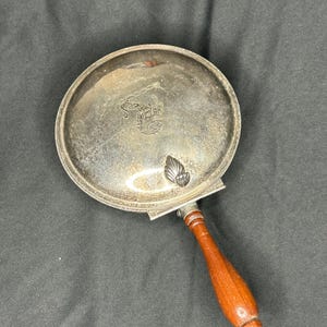 May include: A vintage silver-plated hand mirror with a round, slightly tarnished surface. The mirror features a decorative emblem and a wooden handle with a rich, reddish-brown finish. The handle is attached to the mirror with a silver-colored metal.