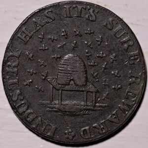 Puede incluir: Una moneda circular de metal marrón oscuro con un relieve de una colmena sobre un soporte en el centro. El texto "INDUSTRY HAS ITS SURE REWARD" está grabado a lo largo del borde exterior, con pequeños símbolos de cruz rodeando la imagen.