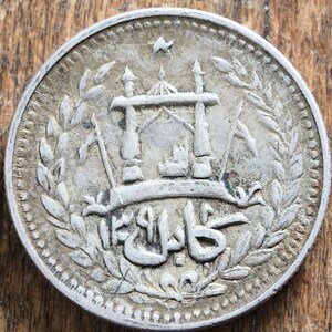 Könnte beinhalten: Eine Silbermünze mit einem Kranz aus Blättern, der ein zentrales Design umgibt. Das Design zeigt ein stilisiertes Gebäude mit einer Kuppel und einem Banner mit dem Wort "Kabul" in arabischer Schrift.
