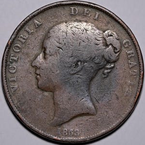 Op de afbeelding: Een zilveren munt met een profiel van een vrouw, met de inscriptie "VICTORIA DEI GRATIA" rond de rand. Het jaartal "1863" is zichtbaar aan de onderkant. De munt heeft een versleten, antieke uitstraling.