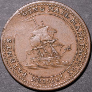May include: A round, copper-colored coin with embossed text and a ship illustration. The text reads "HING NAIL MANUFACTORY PATENTS BRISTOL". The ship has multiple sails and is depicted on the water. The coin has a detailed, aged appearance.