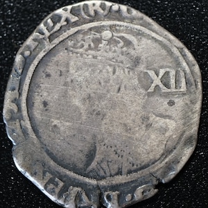 May include: Silver coin with a portrait of a person on one side and the number XII on the other. The coin has a raised rim with text around the edge.