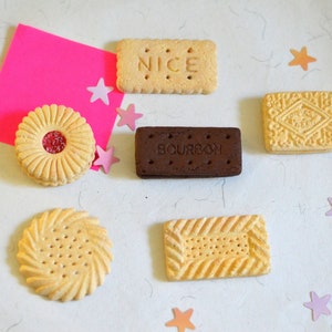 Op de afbeelding: Een assortiment van verschillende smaken biscuits, waaronder een chocoladebiscuit met het label "Bourbon", een biscuit met het label "Nice" en een biscuit met een roze gelei-vulling. De biscuits zijn gerangschikt op een wit oppervlak met roze en oranje sterren.