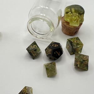 May include: A set of seven green and black dice with gold numbers. The dice are in a glass jar with a cork top. The cork top has a green and black skull on it.