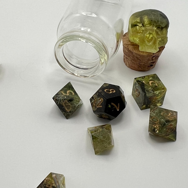May include: A set of seven green and black dice with gold numbers. The dice are in a glass jar with a cork top. The cork top has a green and black skull on it.