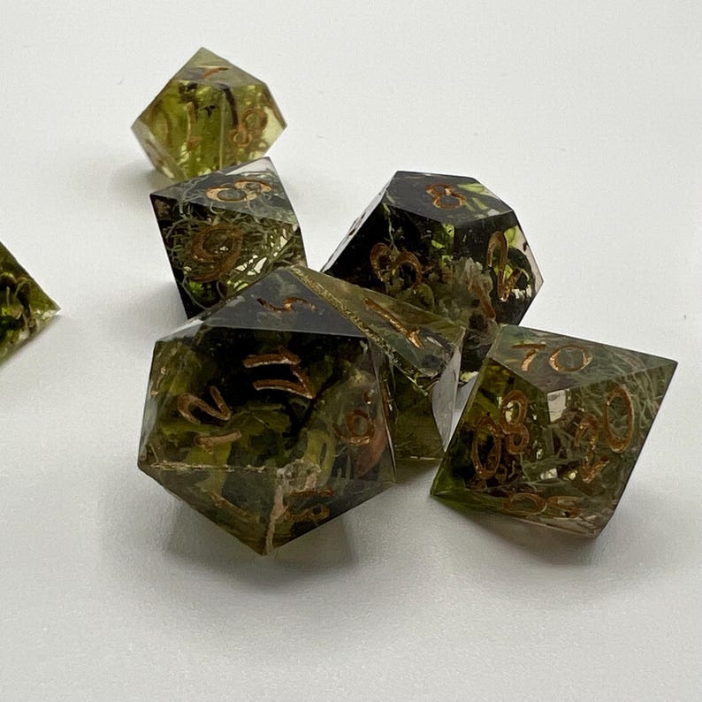 May include: A set of six green and gold dice with a mossy, natural design. The dice are made of clear resin with gold numbers and a green, mossy material embedded inside.
