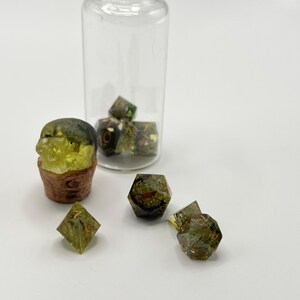 May include: A set of green and gold dice with a skull-shaped die in a glass jar. The dice are made of resin and have a speckled appearance.