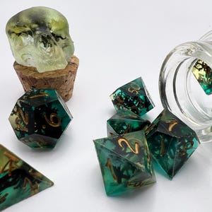 May include: A collection of polyhedral dice with a teal and black design, some scattered from a glass jar. A skull-shaped bottle stopper sits atop a cork. The dice have gold numbering.