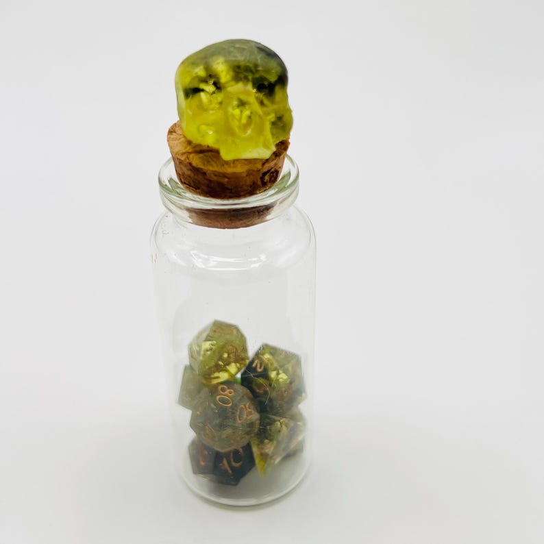 May include: A glass bottle with a cork top shaped like a skull. The bottle contains several green and gold dice.