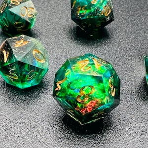 May include: Several polyhedral dice with a green and gold color scheme. The dice are transparent, with gold numbers and internal designs. The dice are arranged on a dark surface, with some in focus and others blurred.
