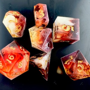 May include: A set of seven polyhedral dice with a marbled effect in shades of red, orange, and white. The numbers are gold. The dice are for tabletop games.