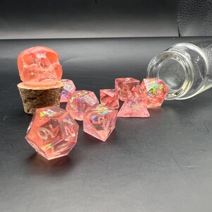 May include: A collection of translucent pink resin dice with iridescent inclusions, scattered near a clear glass bottle. A pink skull-shaped bottle stopper sits atop a cork. The dice are various shapes.