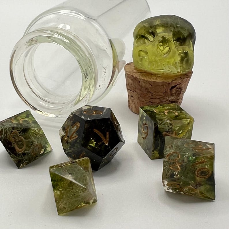 May include: A set of green and black dice with gold accents, including a d20, d4, d6, and d8. The dice are in a glass bottle with a cork stopper shaped like a skull.