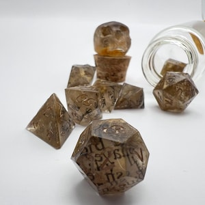 May include: A collection of transparent brown resin dice in various geometric shapes, including a d20, d12, d10, d8, d6, and d4. Some dice have text inside. A glass bottle and a stopper are also present.
