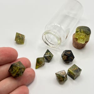 May include: Set of seven green and black dice with gold numbers, including a d4, d6, d8, d10, d%, d12, and d20. The dice are in a glass bottle with a cork stopper and a green skull on top.