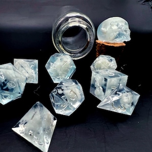 May include: A set of seven translucent blue dice with white numbers and swirls. The dice are on a black surface with a glass jar and a skull-shaped bottle stopper.