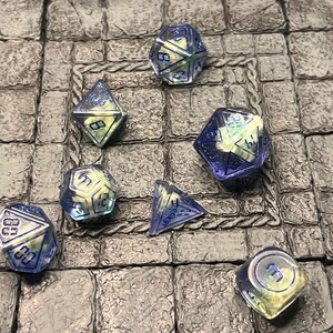 May include: A collection of seven polyhedral dice, each with a translucent blue and yellow color scheme. The dice are scattered on a gray stone surface, with visible numbers on each face. Ideal for tabletop gaming.