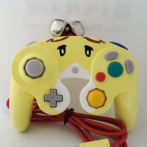 May include: A yellow video game controller featuring a cartoon face with brown hair and black eyes. The controller has a gray directional pad, a yellow C-stick, a green A button, a red B button, and gray X and Y buttons. A red braided cable is attached.