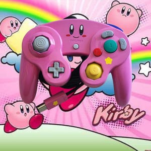 May include: Pink video game controller with a Kirby theme. The controller has a D-pad, analog stick, and colored buttons. The controller has a USB connector. The word "Kirby" is written in a cursive font.