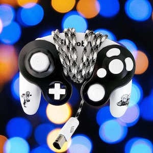以下が含まれることがあります： 白と黒のゲームキューブコントローラー。白いコードと白黒の編組コードが付いています。コントローラーは白黒のデザインで、コードに白いシンボルが付いています。