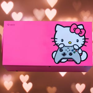 May include: A pink Xbox console with a Hello Kitty design. Hello Kitty is holding a white Xbox controller. The console has the word "XBOX" printed on the front.