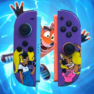 K&ouml;nnte beinhalten: Lila und orangefarbene Gamecontroller-Griffe mit Cartoon-Charakter-Designs. Die Griffe zeigen Bilder von Crash Bandicoot-Charakteren. Der Hintergrund ist ein blaues, elektrisch thematisiertes Design.
