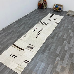 以下が含まれることがあります： 長方形のオフホワイトのパッチワークラグ。長方形と正方形のパッチがいくつかあります。黒と茶色のストライプとアクセントがあります。灰色の木製の床に敷かれています。