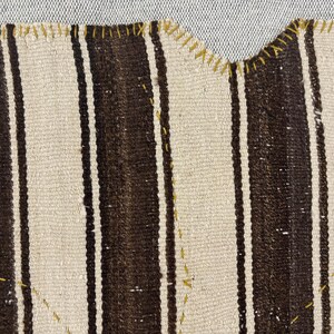 Puede incluir: Un tejido con rayas verticales alternas en marr&oacute;n oscuro y blanco roto. El borde de la tela est&aacute; rematado con una puntada de sobrehilado en hilo amarillo visible, y costuras decorativas amarillas crean patrones geom&eacute;tricos en la superficie.
