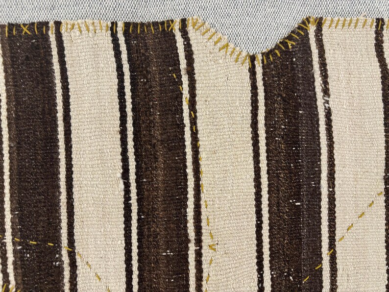 Puede incluir: Un tejido con rayas verticales alternas en marr&oacute;n oscuro y blanco roto. El borde de la tela est&aacute; rematado con una puntada de sobrehilado en hilo amarillo visible, y costuras decorativas amarillas crean patrones geom&eacute;tricos en la superficie.